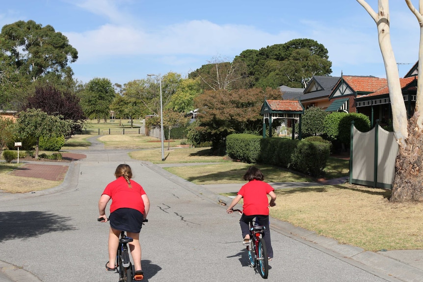 Two girls in red shirts riding bikes down a street, lined with houses and trees.