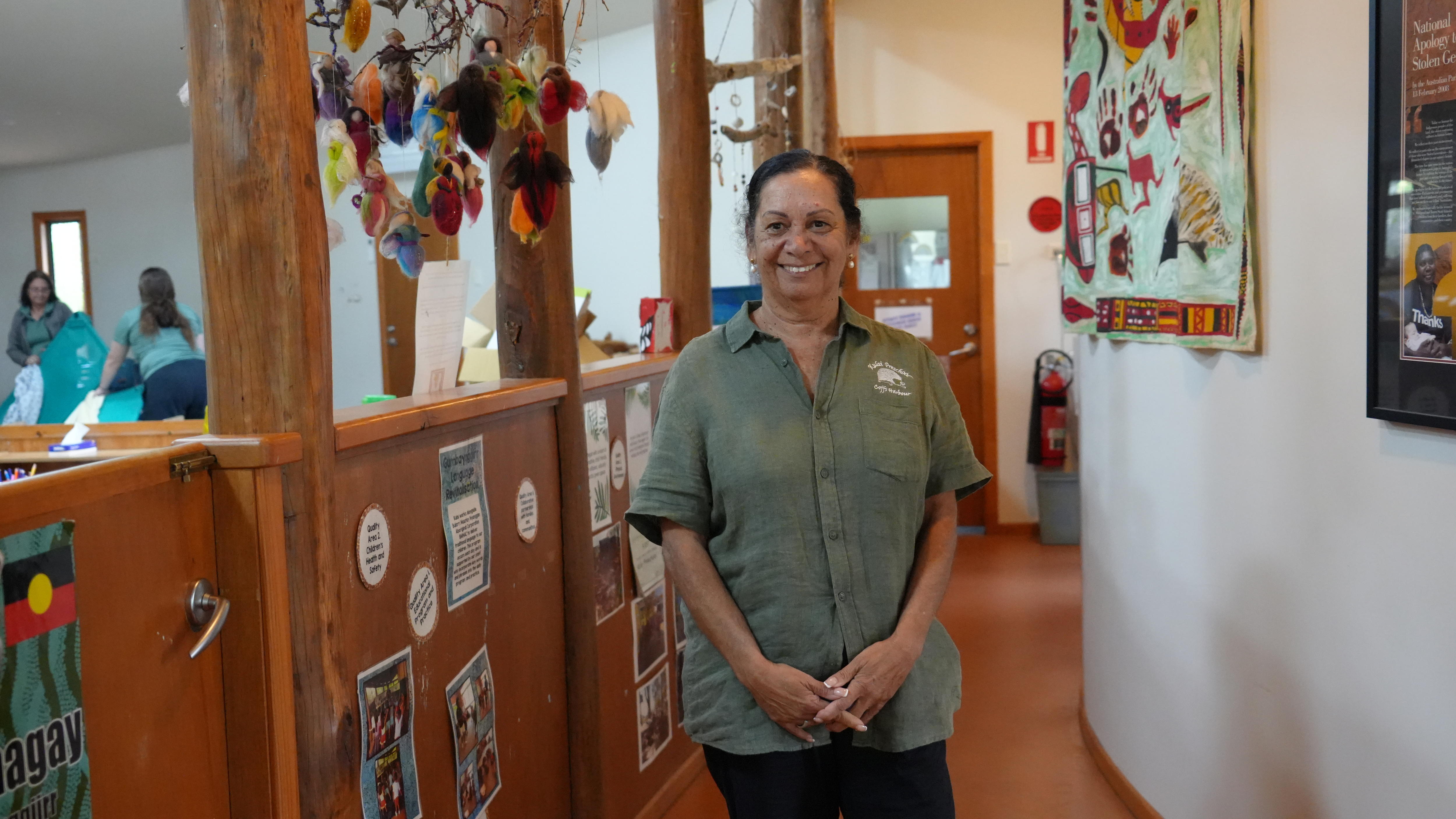 An Aboriginal woman in a colourful preschool space wearing a khaki green Kulai shirt