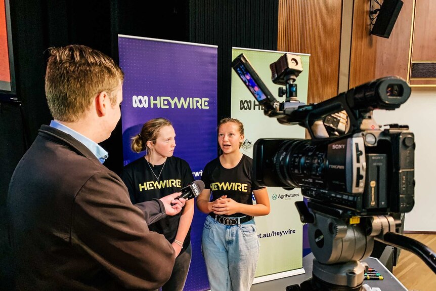 Two young people stand in front of a camera, interviewed by a journalist with a microphone