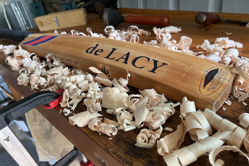 A cricket bat sits on a workbench surrounded by wood shavings.