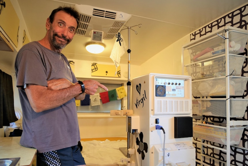 A man standing in a caravan with pointing at a dialysis machine.