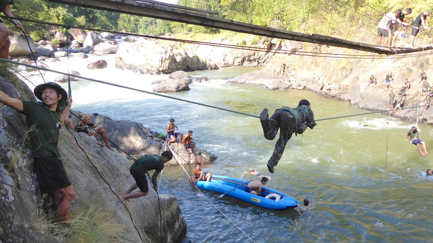 A person crawls along a rope suspended above a river, as others abseil and swim below.