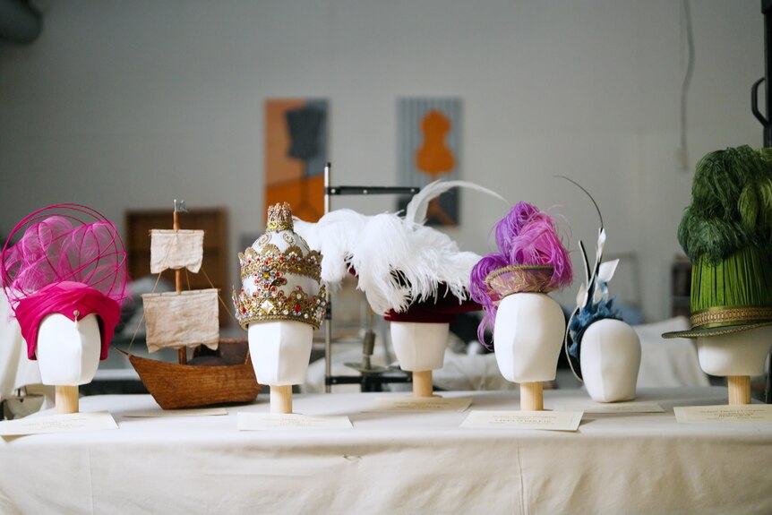 Six intricate hats of different vibrant colours sit on a table with a white tablecloth. 
