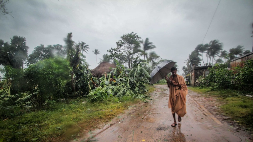 A man in an orange sari tried to hold an umbrella as heavy rains fall, trees and vegetation uprooted around him.