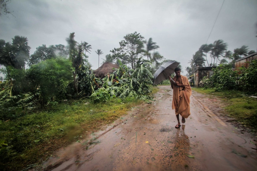 A man in an orange sari tried to hold an umbrella as heavy rains fall, trees and vegetation uprooted around him.