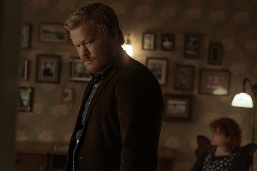 A blonde bearded man wears brown coat and stands looking down in warmly lit floral interior wall with hanging picture frames.