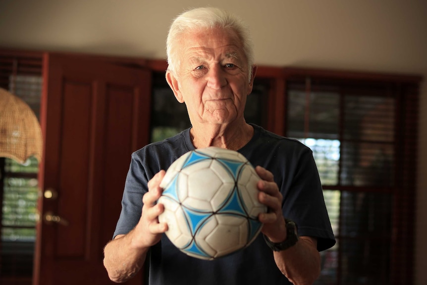 Peter Webster holds a blue and white soccer ball while staring at the camera inside his home.