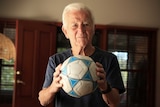 Peter Webster holds a blue and white soccer ball while staring at the camera inside his home.