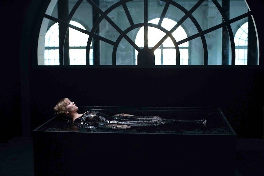 A woman lies down in a large container of water, in front of an intricate window