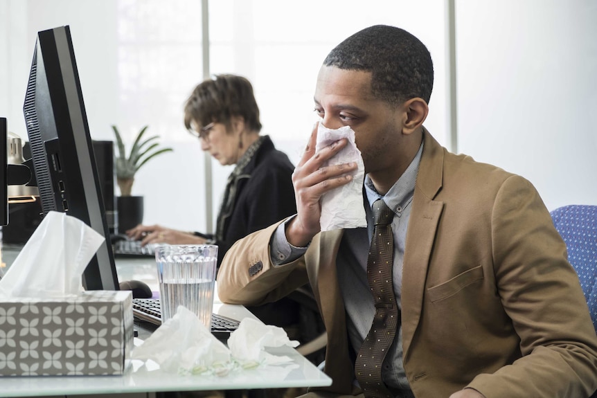 A man sits at a desk looking at a computer while blowing his nose.