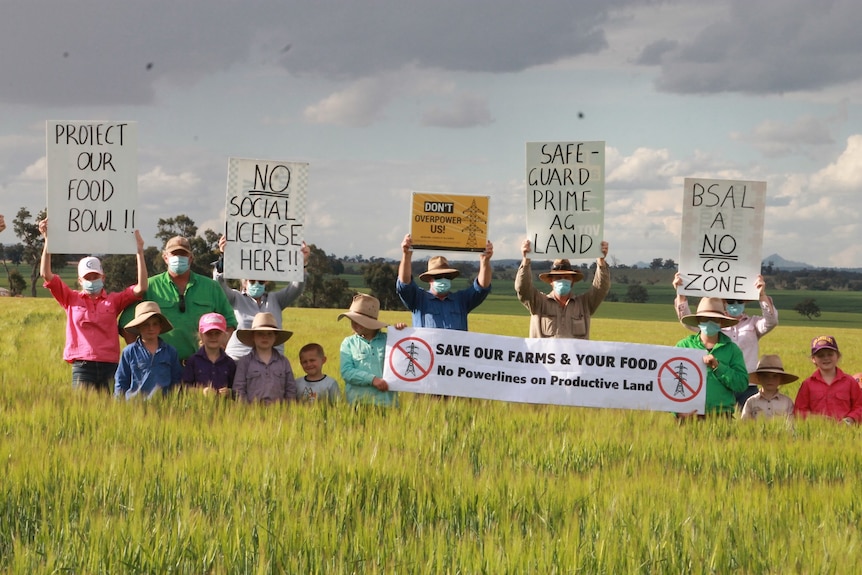 A group of farmers stand in a paddock holding up placards reading 'safe guard prime ag land', 'we will fight for our land'...