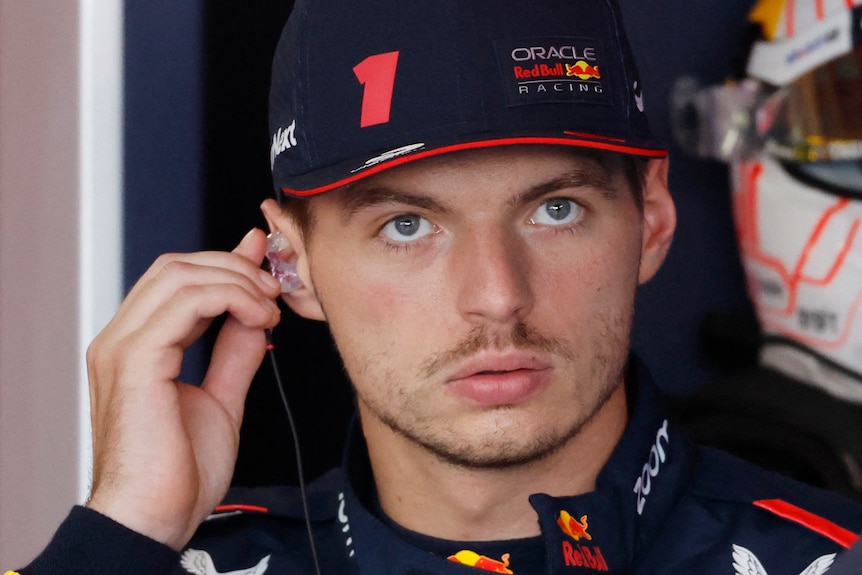 An F1 driver in a blue shirt and blue cap, putting an earplug into one ear, looking out from his garage