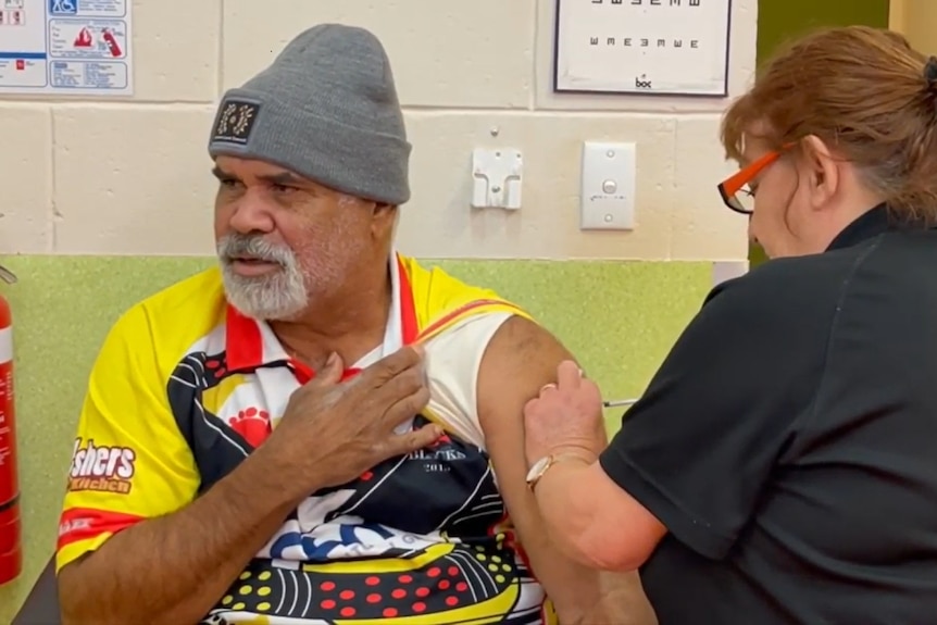 Sammy Wilson, an Indigenous man, holds his shirt sleeve up while receiving a vaccination in his left arm.