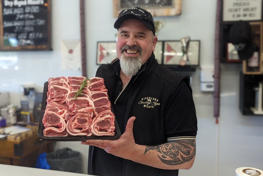 A white smiling man with a beard wearing a black cap holds up a tray of lamb loin chops in his butcher shlop.