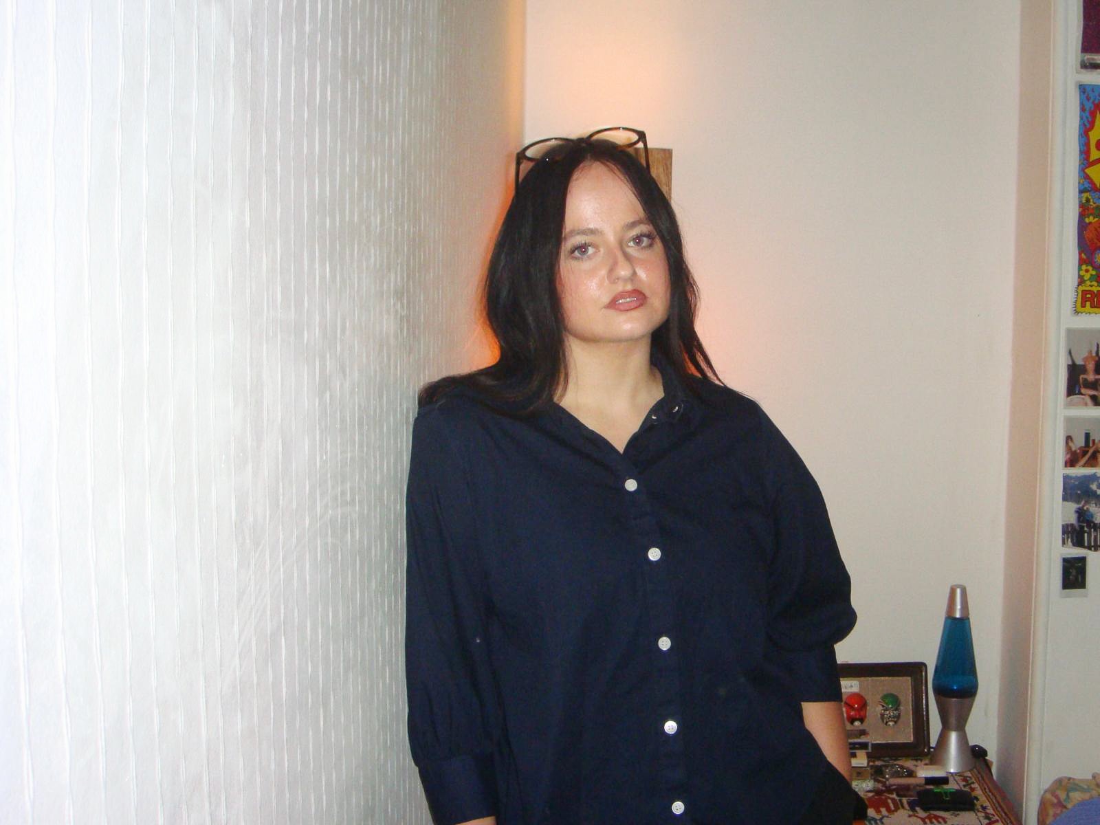 A young woman with brown hair leans against a wall, she wears a navy blue oversized shirt and glasses on her head