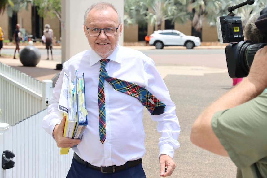 A middle aged man with grey hair leaving a building, holding folders, with camera operators surrounding him