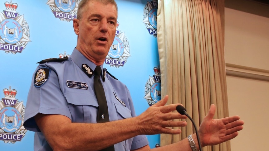 Karl O'Callaghan stands at a podium indoors talking in a police uniform with his arms outstretched.