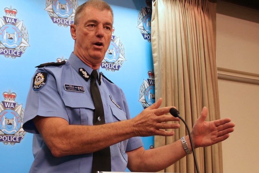 Karl O'Callaghan stands at a podium indoors talking in a police uniform with his arms outstretched.