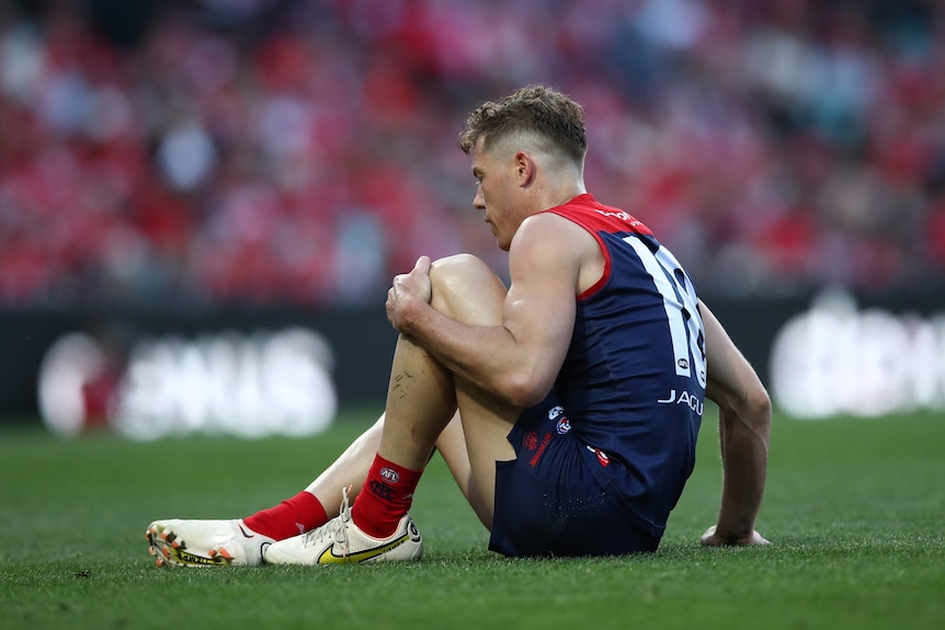A Melbourne AFL player sits on the ground during a match holding on to his knee.