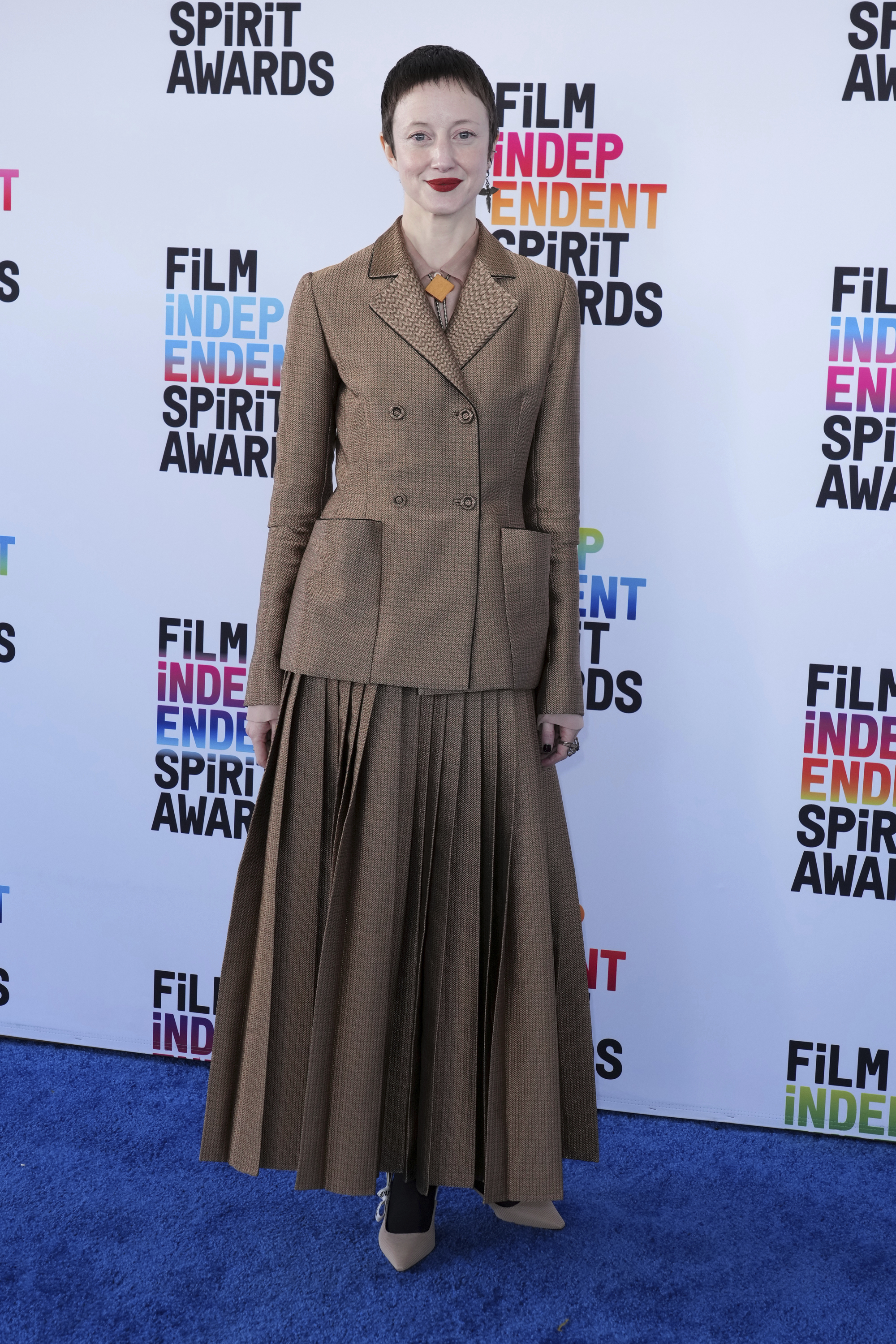 Andrea Riseborough wearing a brown tweed-like blazer with a matching ankle-length pleated skirt 