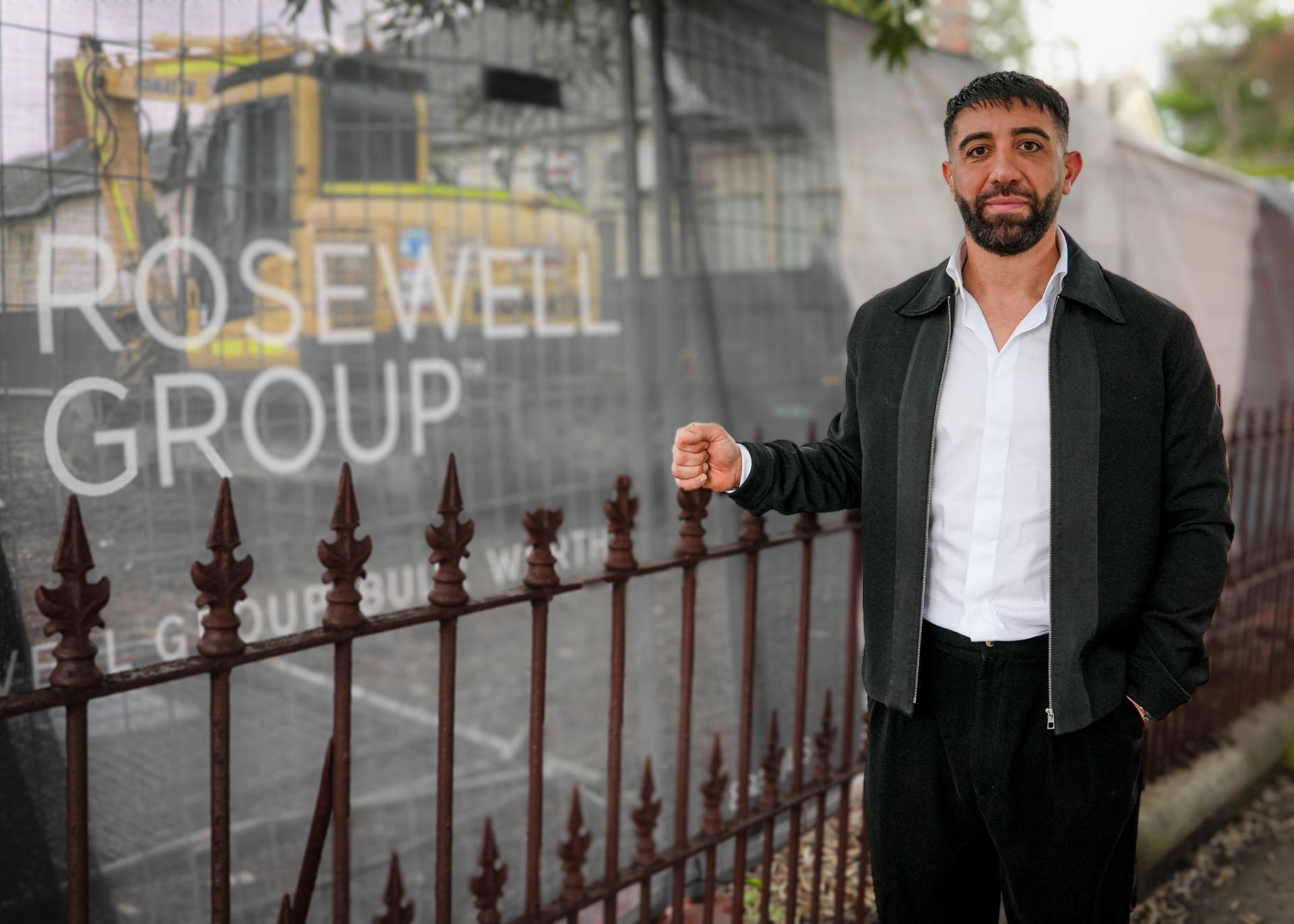 A dark-haired, bearded man stands next to a construction site.