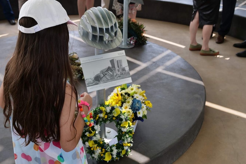 A young girl reads a plaque at a memorial with floral wreaths at the base.
