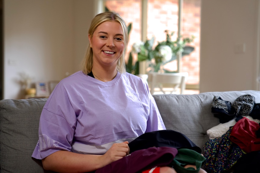 Netballer Eleanor Cardwell sits on a couch, surrounded by sports bras. She's wearing a purple t-shirt and is smiling.