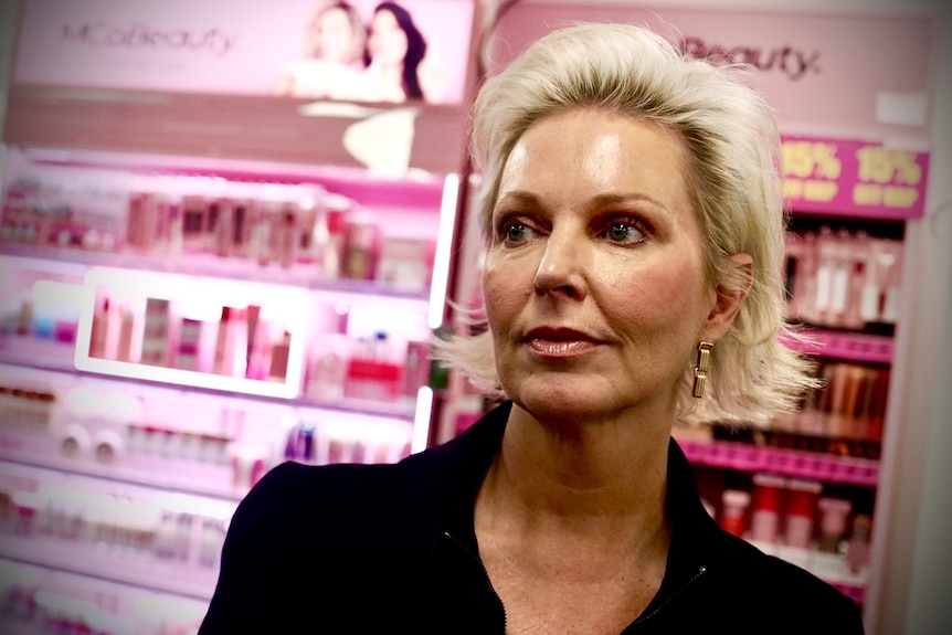 A woman with short blonde hair looks off to the side standing in front of a pink cosmetics display.