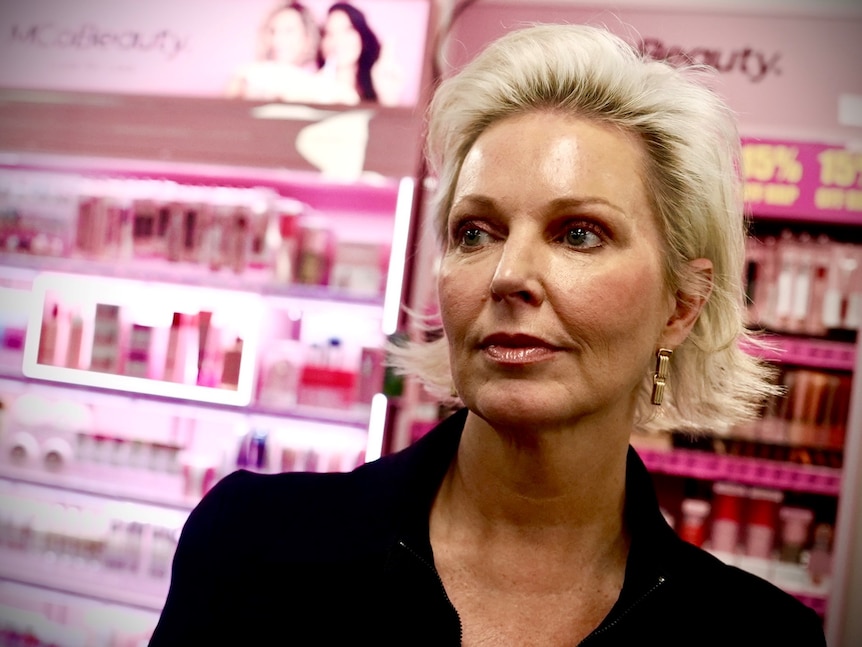 A woman with short blonde hair looks off to the side standing in front of a pink cosmetics display.