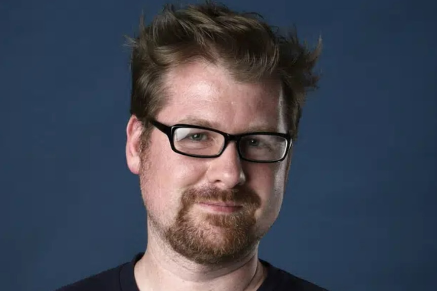 A young white man with a goatee in a black tshirt on a blue backdrop
