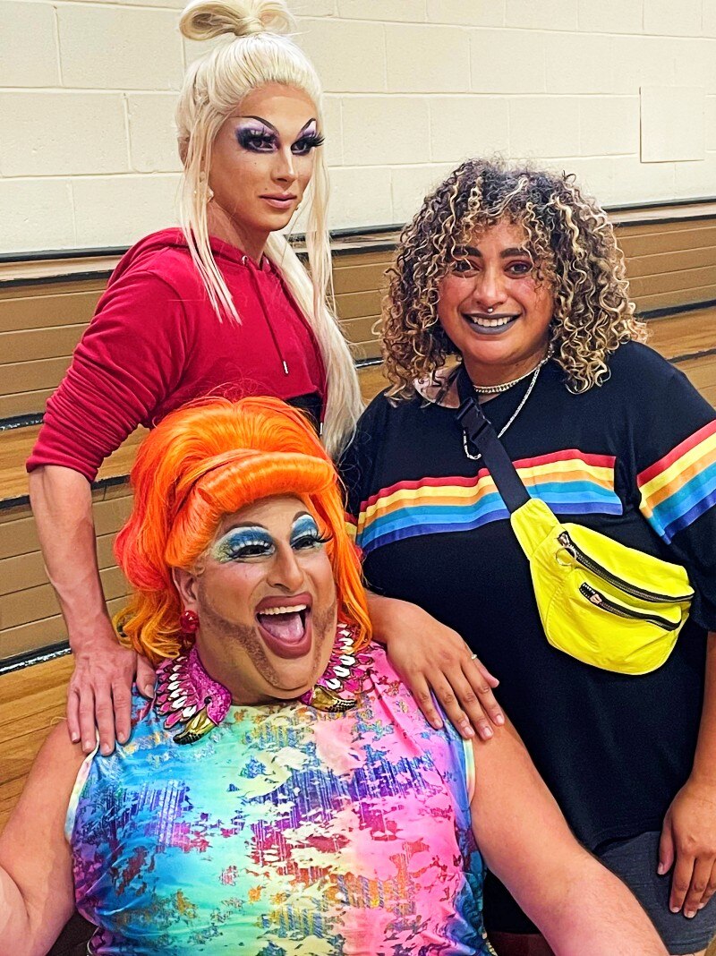 Three people in bright coloured clothes and makeup on the edge of a basketball court