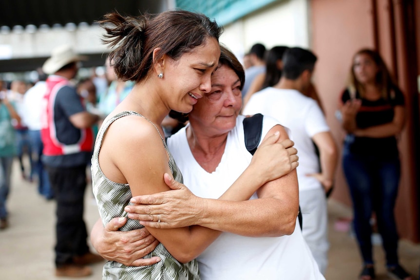 Two women hug each other with tearful faces with a group of others standing in the background