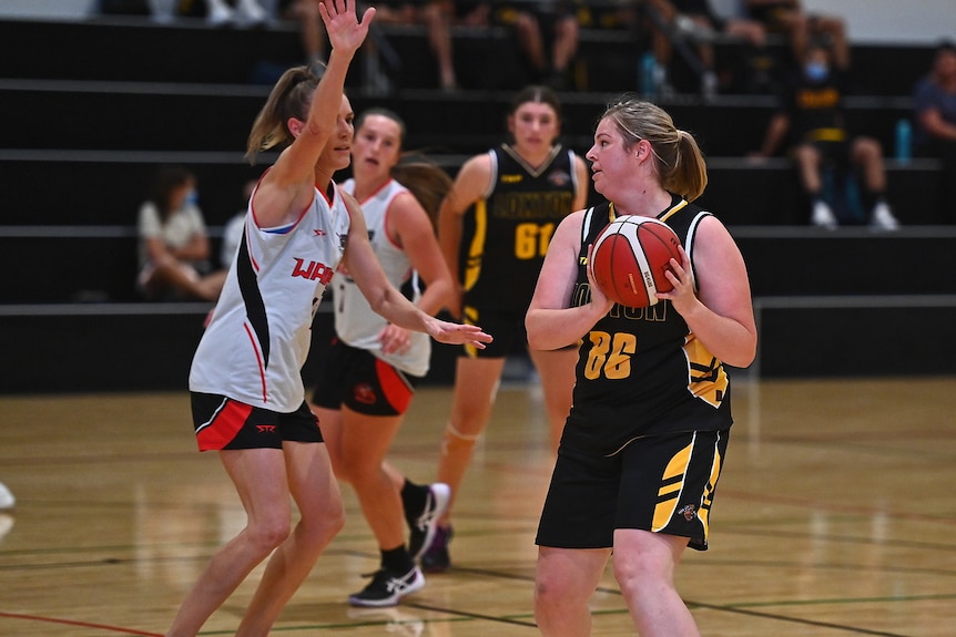 A woman wearing a black and yellow basketball uniform holding a ball in front of a woman wearing a white uniform. 