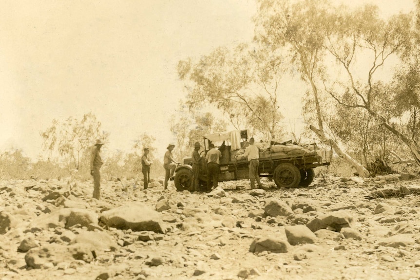 A few men stand around a mail truck it is parked on the rocky, uneven ground of a dried riverbed. The old photo is sepia-toned.