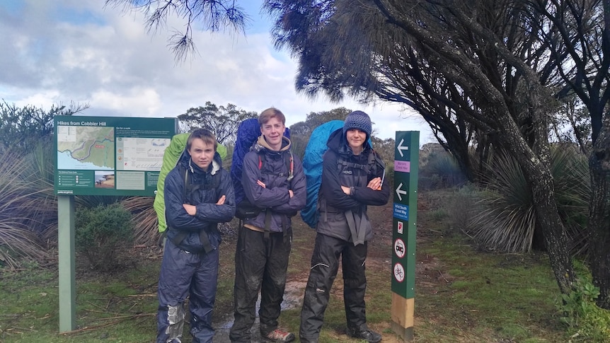 Three boys with large camping packs on their backs stand in front of a sign