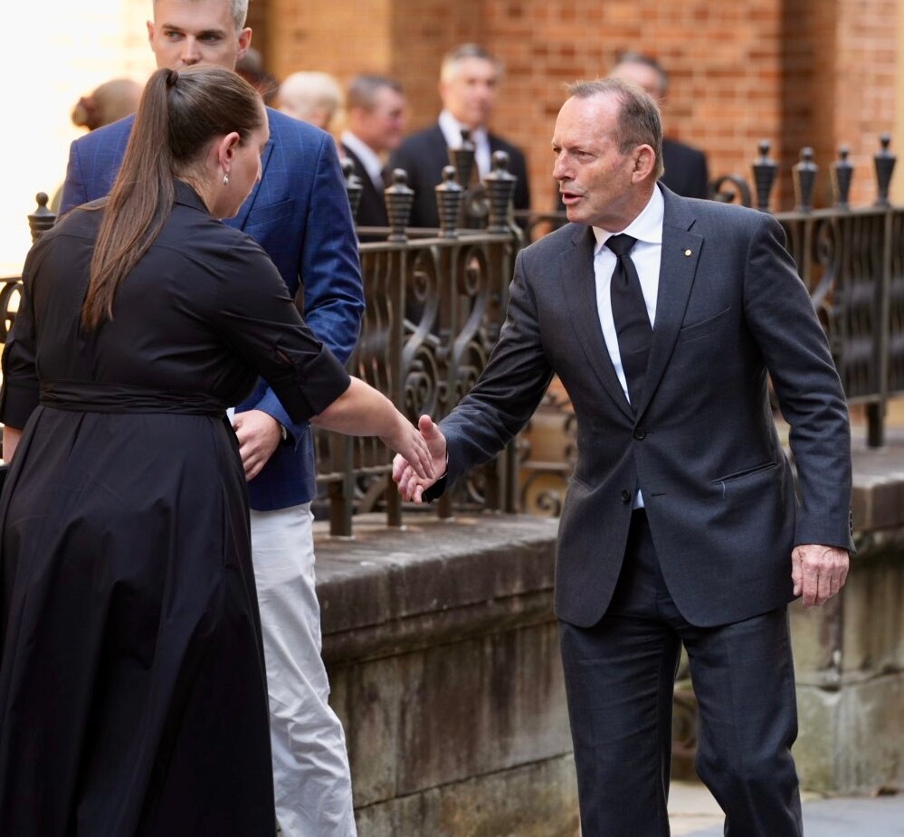 Con traje oscuro y corbata, Tony Abbott le da la mano a una mujer con un vestido negro.