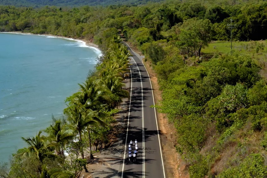 Bike riders on a straight stretch of road with the ocean on their left and rainforest on their right