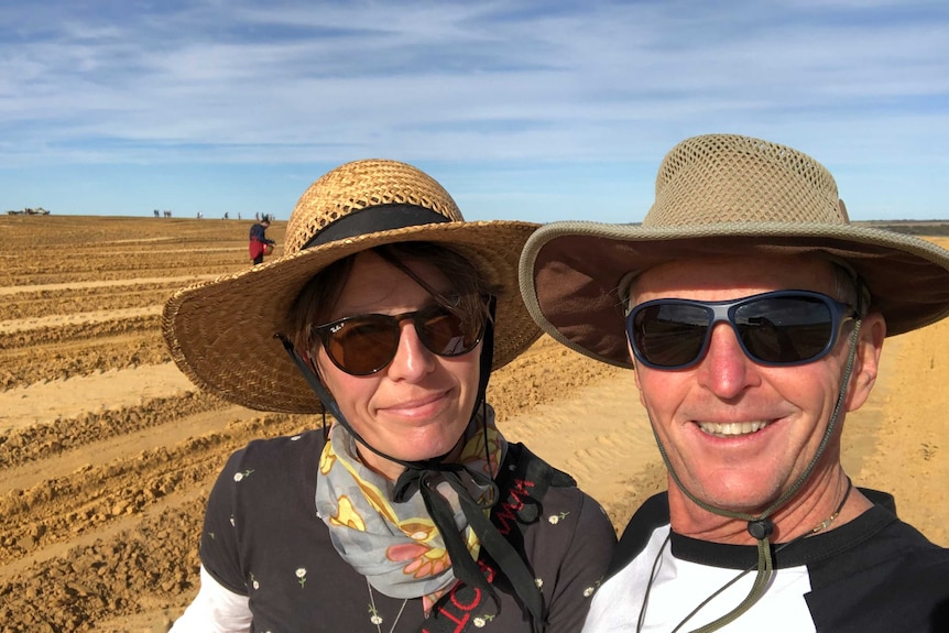 A man and a woman wearing hats and sunglasses smile in a selfie as they stand in a field of dirt.