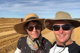 A man and a woman wearing hats and sunglasses smile in a selfie as they stand in a field of dirt.