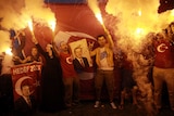 Group of people standing in front of Turkish flag hold flares and pictures of Tayyip Erdogan