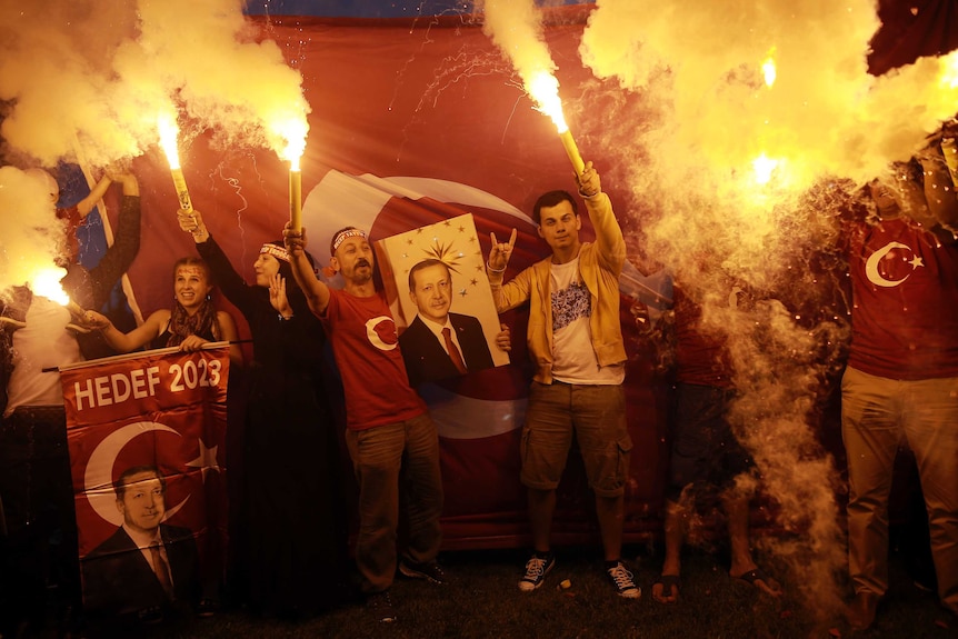 Group of people standing in front of Turkish flag hold flares and pictures of Tayyip Erdogan