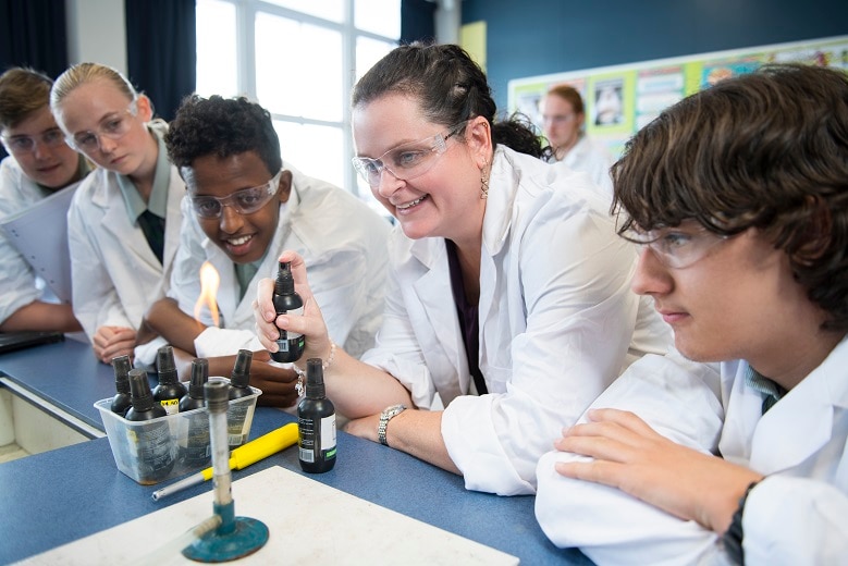 Woman surrounded by four children in lab coats sprays a lit bunsen burner with a chemical