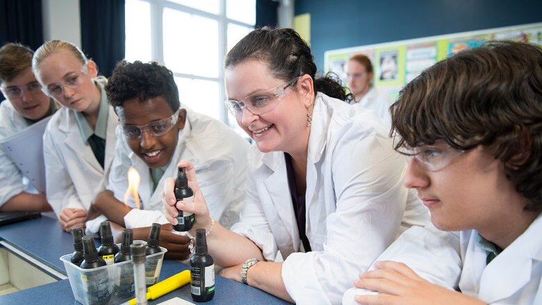 Woman surrounded by four children in lab coats sprays a lit bunsen burner with a chemical
