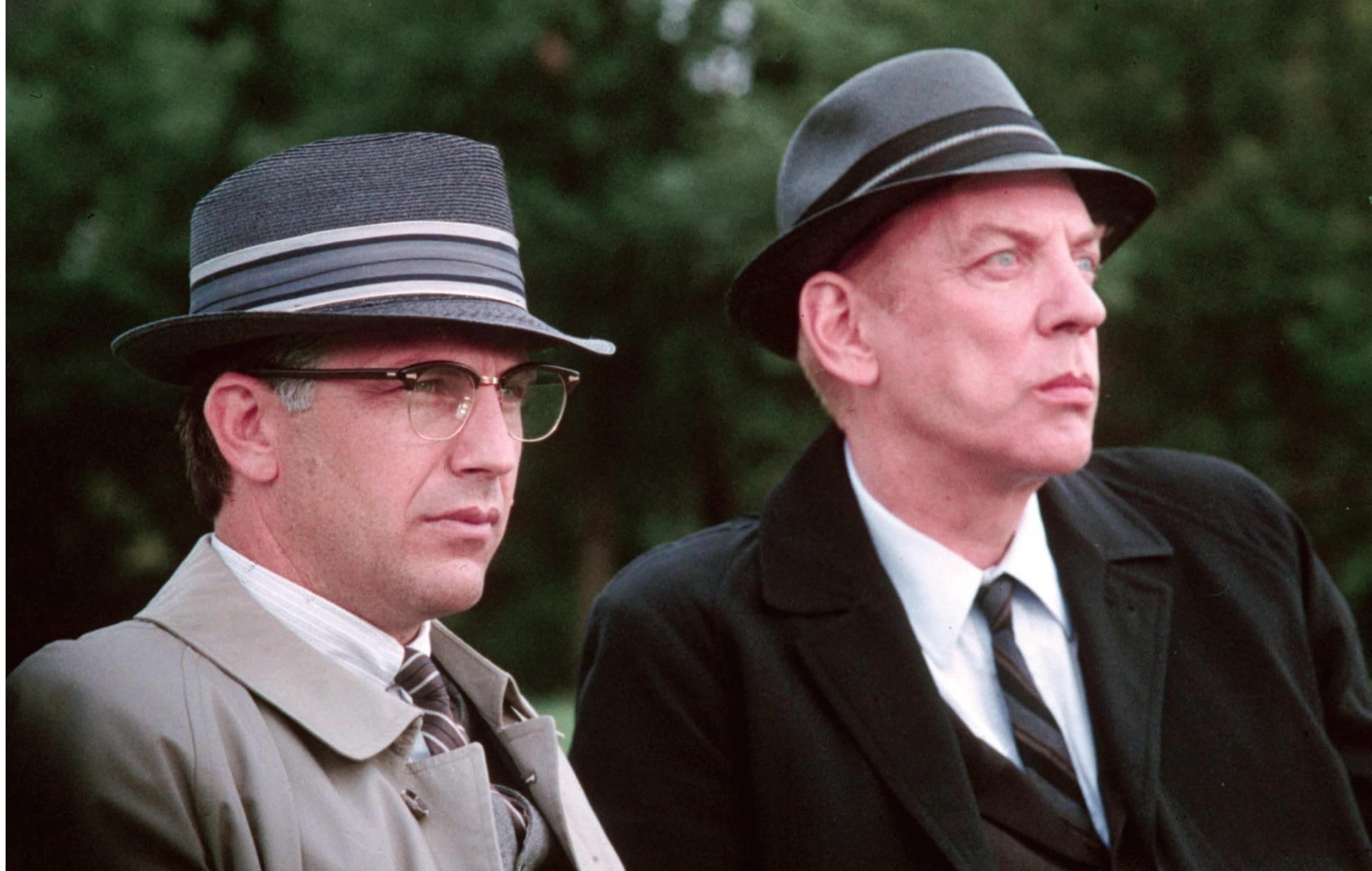 Kevin Coster and Donald Sutherland in the 1991 film JFK sitting on a bench with suit and hats on