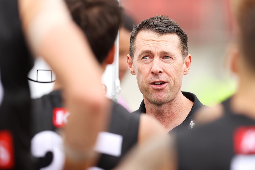 The camera looks through a crowd of AFL players to focus on a coach who is standing, addressing his team during a match.