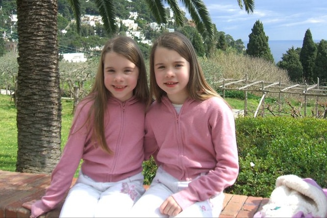 Two twins, aged nine, sit on a brick wall in a garden, wearing same clothes, smile at the camera.