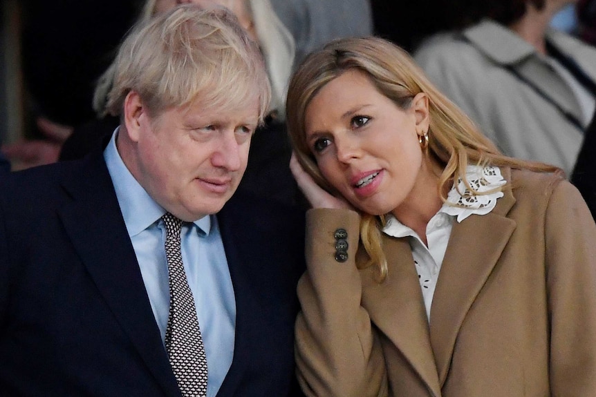 Boris Johnson in a suit and tie looks off camera as Carrie Symonds leans in towards him