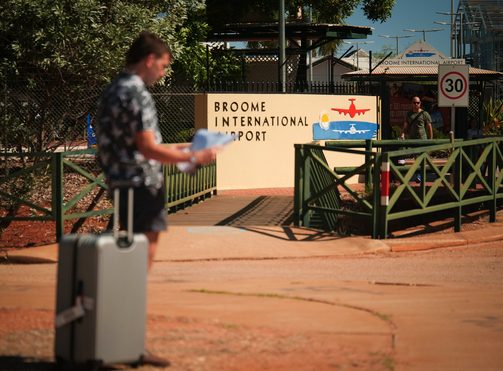 A man stands near a suitcase, looking at a map in front of a sign that says "Broome International Airport".