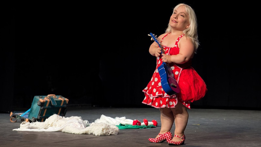 Kiruna Stamell stands on stage in a red dress and holds a blue ukulele. An open suitcase is seen behind her.