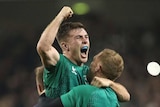 A man wearing a green rugby top clenches his fist and screams while being held up by another man